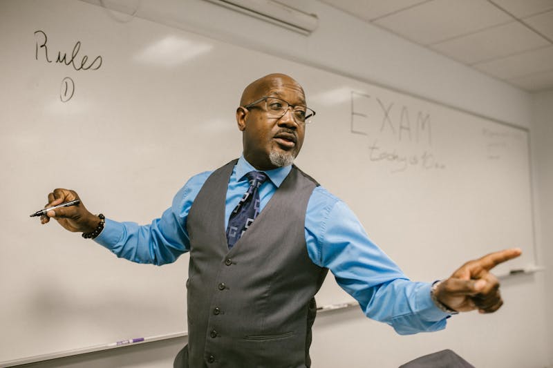 An examiner standing in front of a whiteboard with the word 'EXAM' written on it, briefing candidates before a high-stakes assessment.